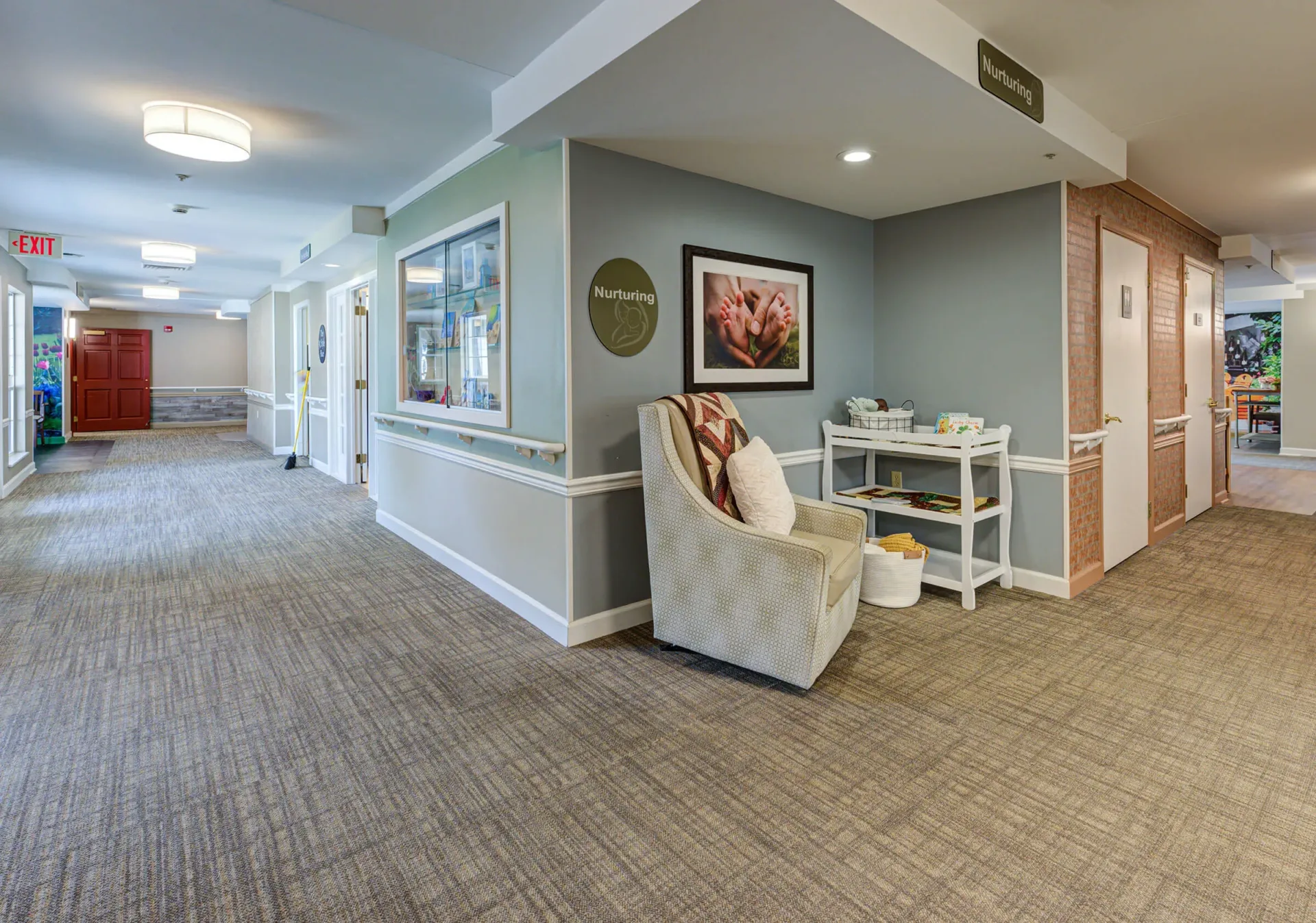 A hallway in Arden Courts Memory Care features beige carpet, light-colored walls, a cushioned chair, a small shelf with items, and several doors leading to rooms.