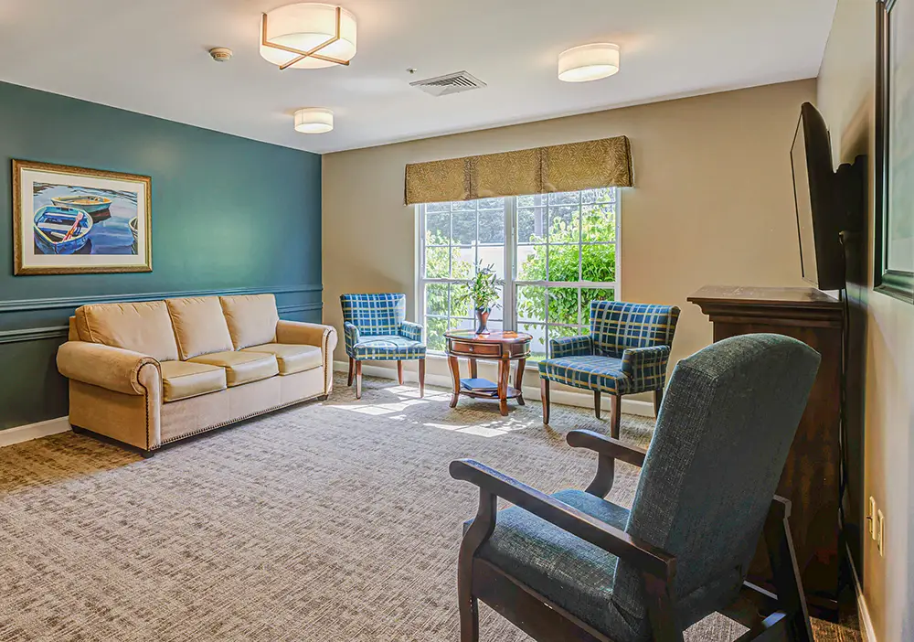 A living room at Arden Courts Memory Care features a beige sofa, two blue plaid chairs, a dark armchair, wooden table, wall art, carpeted floor, and a large window filling the space with natural light.