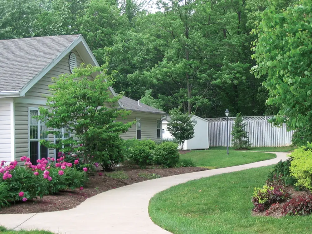 A concrete sidewalk curves through a landscaped yard with green grass, bushes, and flowering plants at Arden Courts Memory Care, next to a light-colored house.