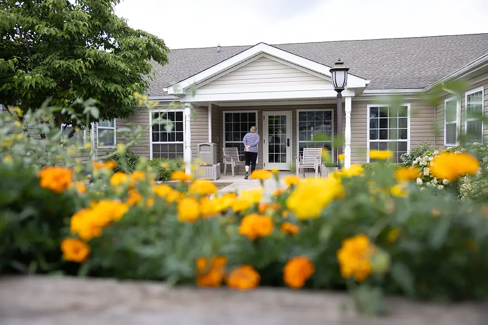 A person walks toward the entrance of Arden Courts Memory Care, a single-story building with yellow flowers in the foreground.