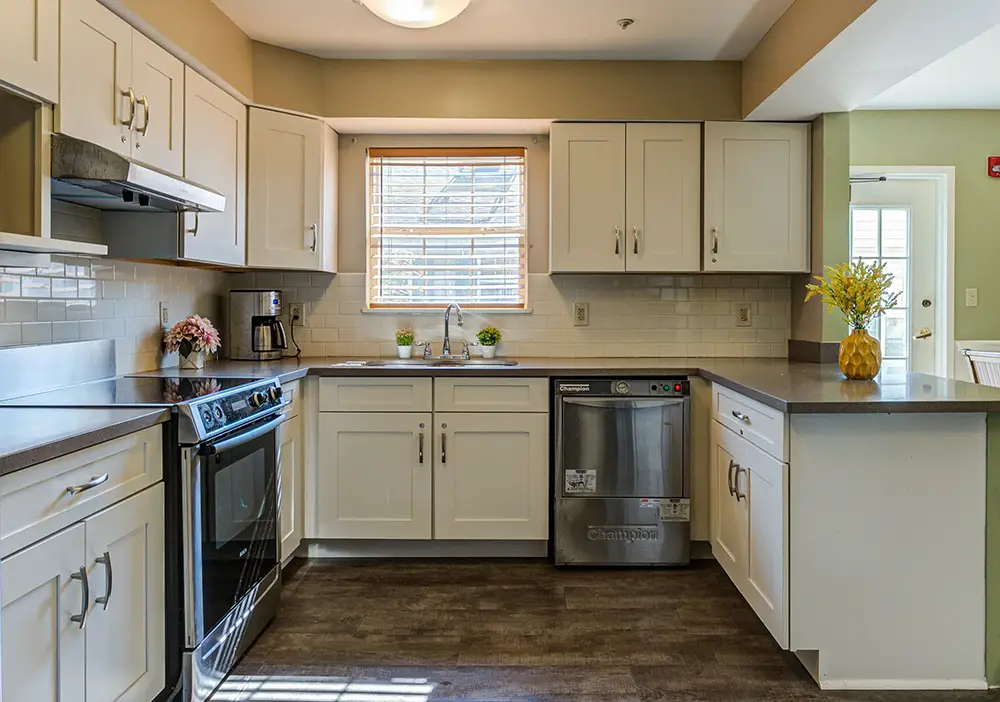Modern kitchen with white cabinets, stainless steel appliances, a window over the sink, and decorative plants and flowers on the counters, inspired by the bright and welcoming style found at Arden Courts Memory Care.