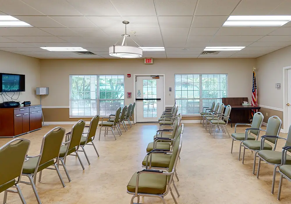A brightly lit room at Arden Courts Memory Care features rows of green chairs arranged in a semi-circle, a TV on the wall, large windows, and an American flag in the corner.