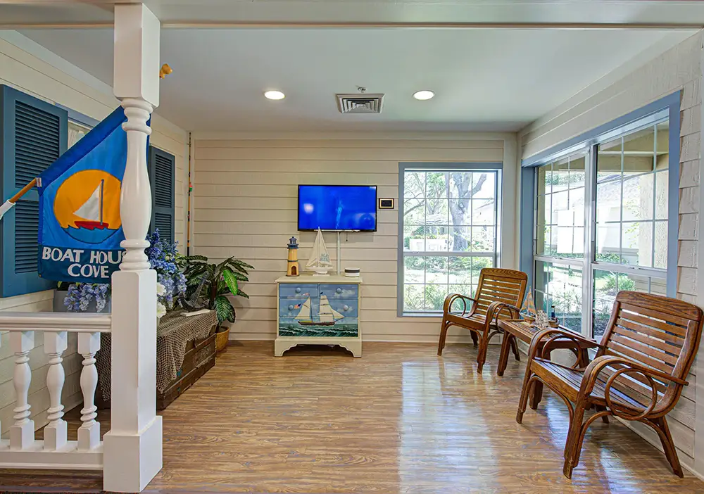 A bright room at Arden Courts Memory Care features nautical decor, a blue flag, wooden benches, a TV on the wall, and large windows letting in natural light.
