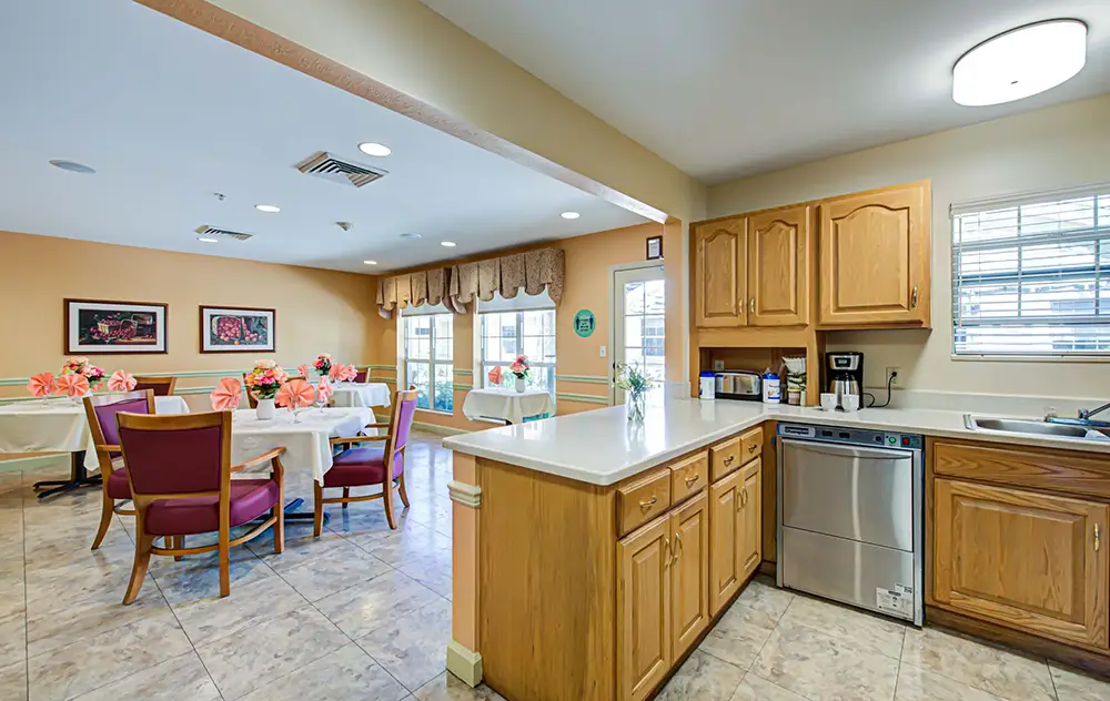 A bright dining area at Arden Courts Memory Care features round tables with flowers, next to a kitchen with wooden cabinets, white countertops, a dishwasher, and a sink.