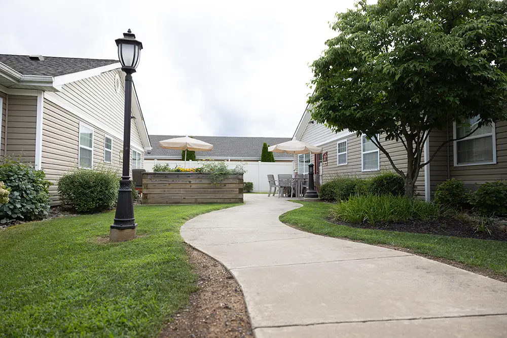 A curved concrete walkway runs between two beige buildings at Arden Courts Memory Care, with a streetlamp, green lawn, trees, and patio tables with umbrellas in the background.
