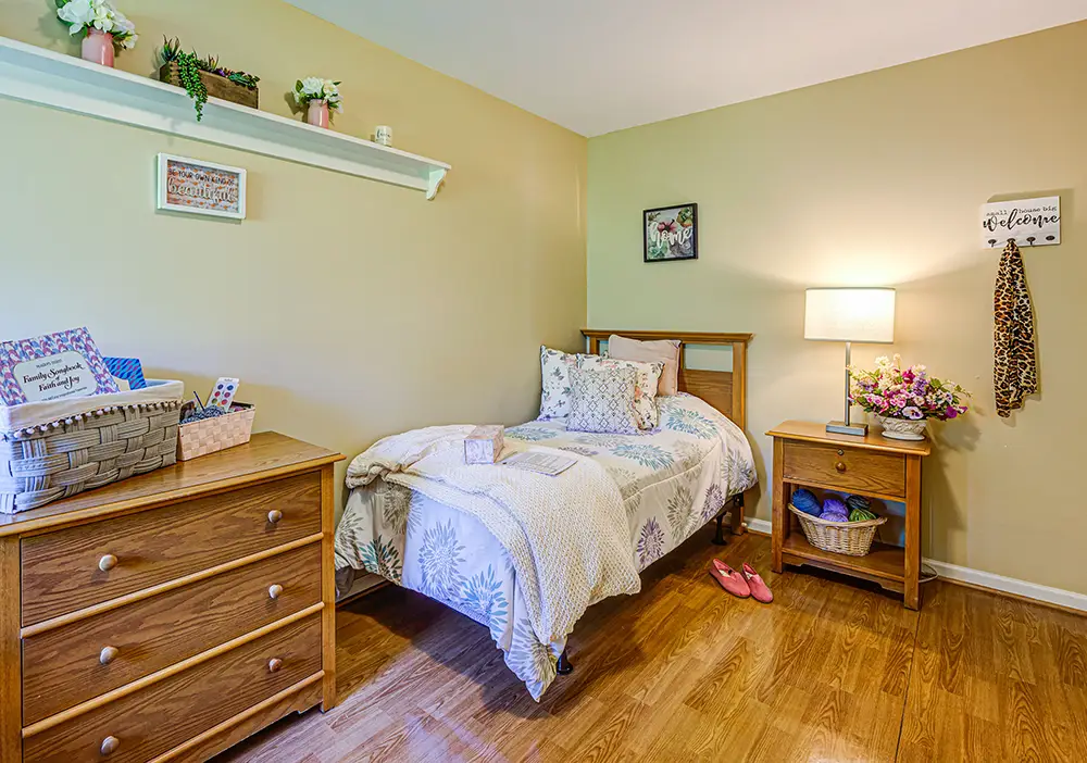 A neatly made single bed in a cozy Arden Courts Memory Care bedroom with a wooden dresser, nightstand, lamp, floral decor, and hardwood floor.