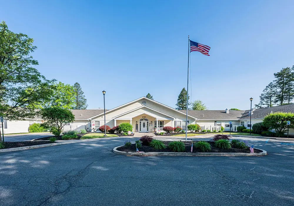 A single-story building with a gabled roof sits behind a circular driveway at Arden Courts in Allentown, with an American flag on a pole and landscaping on a clear, sunny day—designed for comfortable memory care.