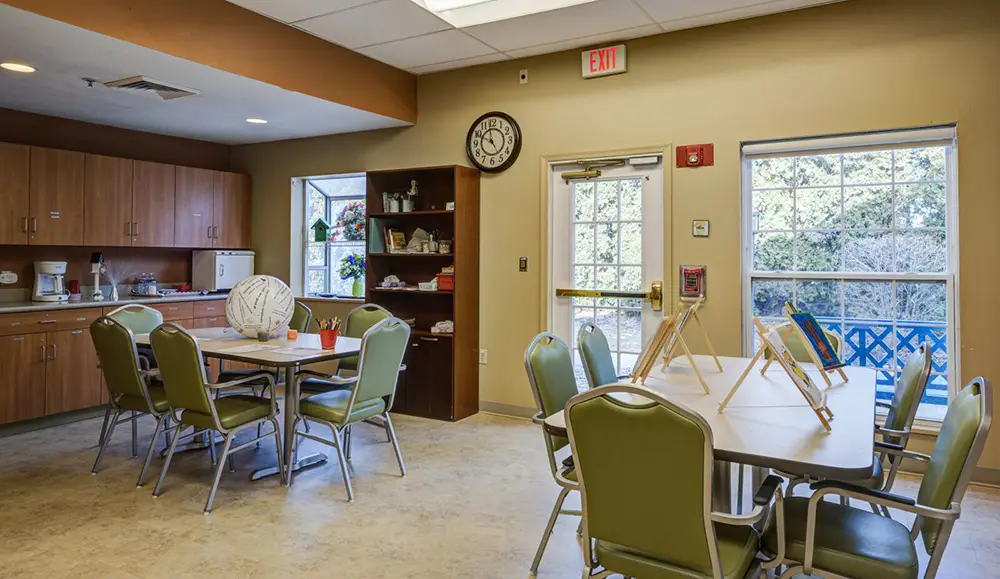 Allentown11 A community room with green chairs around tables, art easels, cabinets, a clock on the wall, and windows letting in natural light.