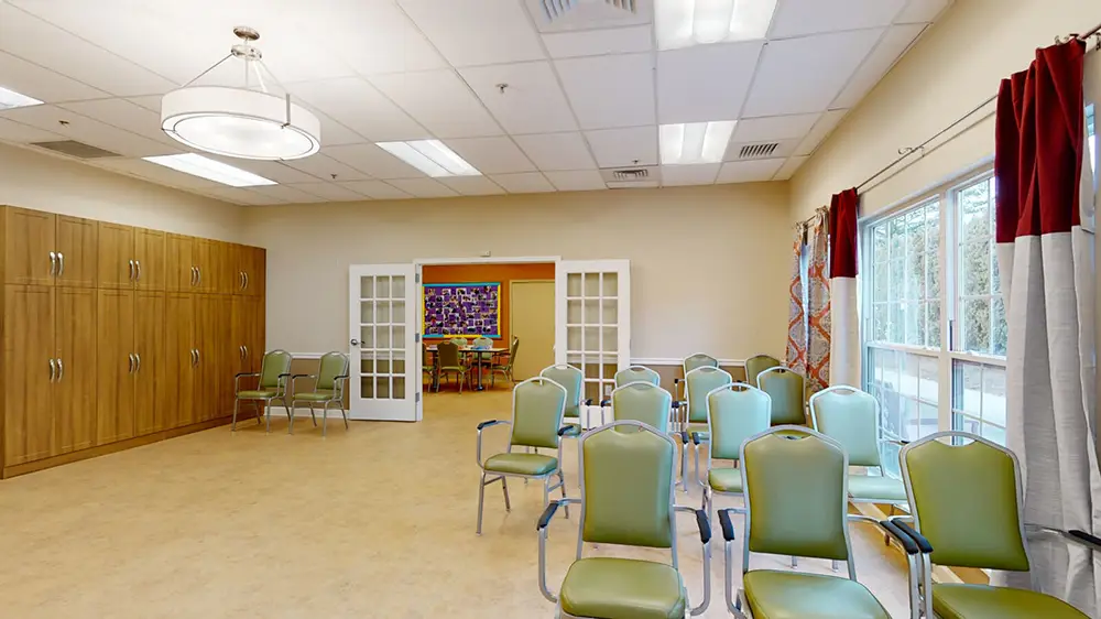 Allentown12 Small meeting room with green chairs arranged in rows, wooden cabinets on the left, and double doors leading to a colorful room with a table and chairs in the background.