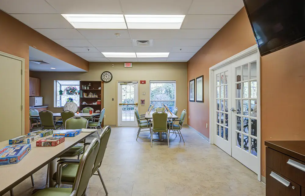 Allentown13 A well-lit activity room with tables, chairs, board games, a wall clock, and double glass doors leading to an outdoor area.