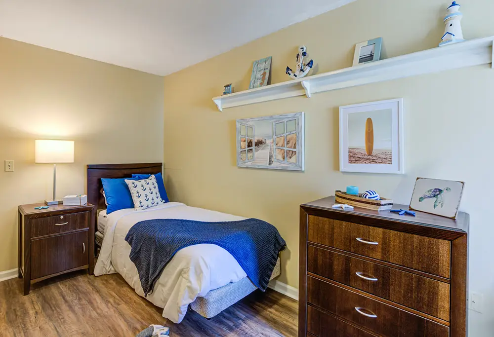 Allentown15 A neatly made twin bed with blue and white bedding beside a wooden nightstand and a dresser, in a room decorated with nautical-themed decor and wall art.