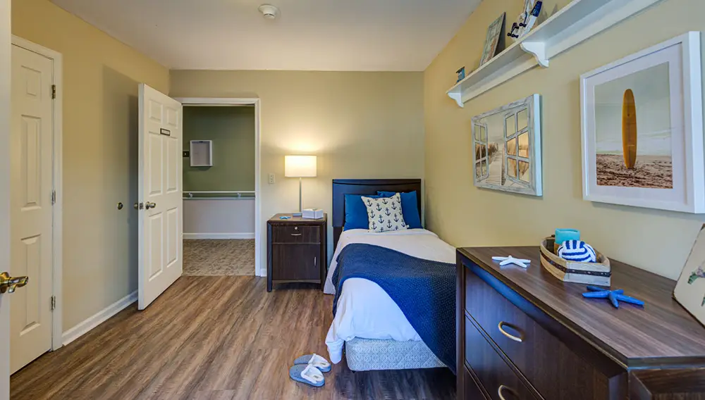 Allentown17 A neatly made single bed with blue and white bedding sits beside a nightstand in a small, well-lit bedroom with wood flooring and beach-themed decor.