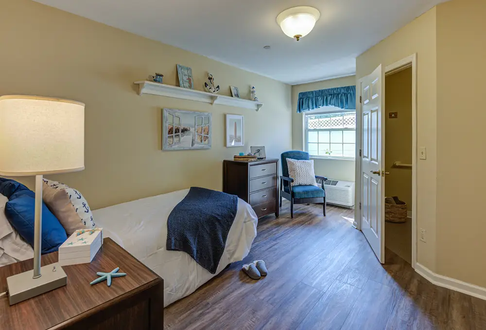 Allentown18 A neatly furnished bedroom with a single bed, dresser, armchair by a window, and a small table lamp, featuring wooden floors and beige walls.
