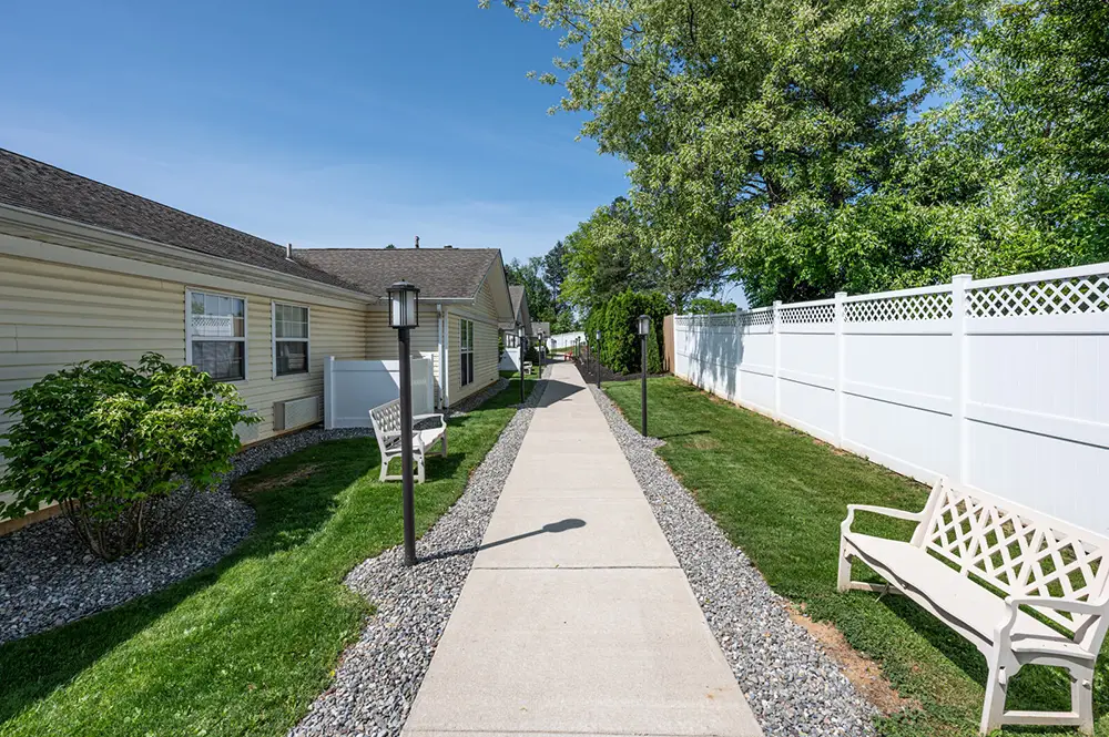 Allentown2 A concrete walkway runs between a beige building and a white fence, with benches, shrubs, and trees along the path on a sunny day.