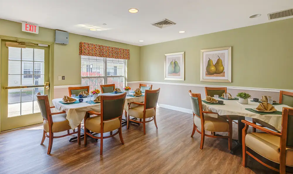 Allentown20 A dining room with two round tables set for meals, surrounded by wooden chairs. The room has green walls, framed pear artwork, and a window with a patterned valance.