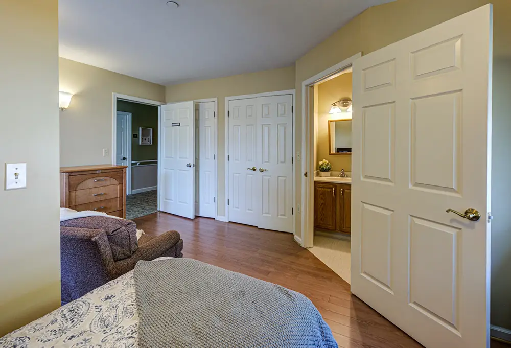 Allentown21 A bedroom with wood floors, a bed, an armchair, a dresser, three closed closet doors, and an open door leading to a bathroom with a vanity and mirror.