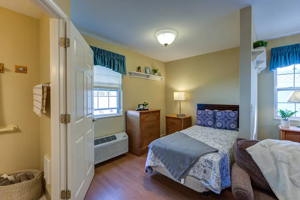 Allentown25 A neatly arranged bedroom with a double bed, wooden furniture, an armchair, two windows with blue curtains, and a laundry basket near a partially open door.