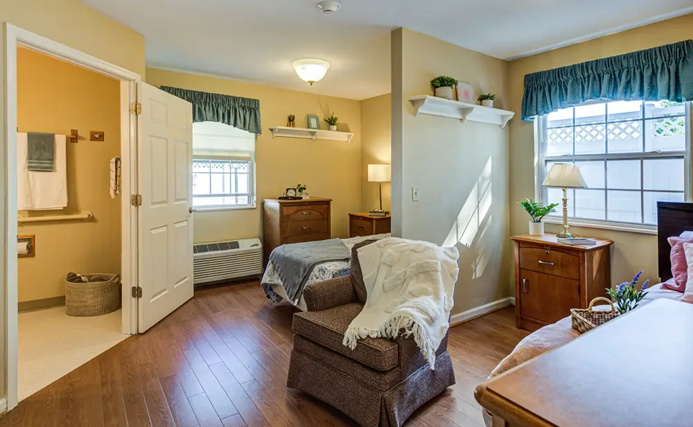 Allentown26 A tidy, well-lit bedroom with two beds, wooden furniture, a large window with valance curtains, shelves with plants, and an open door leading to an accessible bathroom.