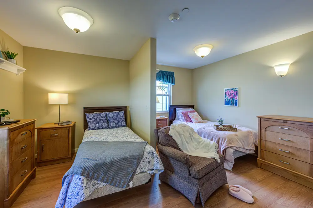 Allentown27 A shared bedroom with two single beds, wood furniture, a chair, and soft lighting; a window and framed art are visible on the wall.