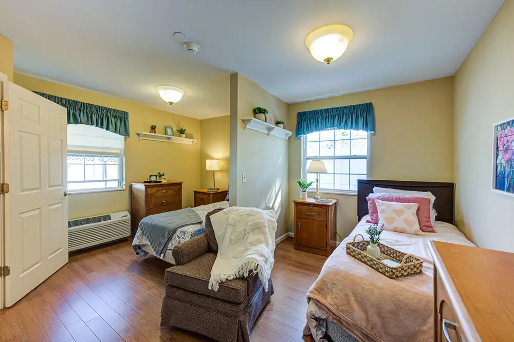 Allentown28 A tidy bedroom with two single beds, a cushioned armchair, wooden nightstands, dressers, potted plants, and large windows with green curtains letting in natural light.