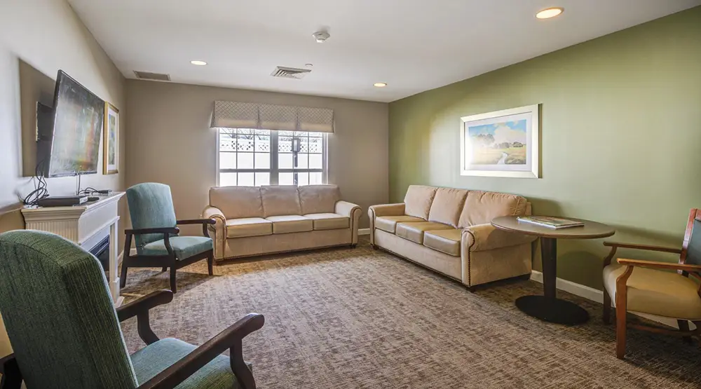 Allentown29 Small living room with two beige sofas, green armchair, round table with magazine, wall art, and a TV mounted near a window with patterned valance.