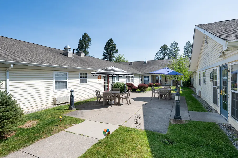Allentown3 Outdoor courtyard at a residential facility with patio tables, chairs, umbrellas, and surrounding lawn, bordered by one-story beige buildings under a clear sky.