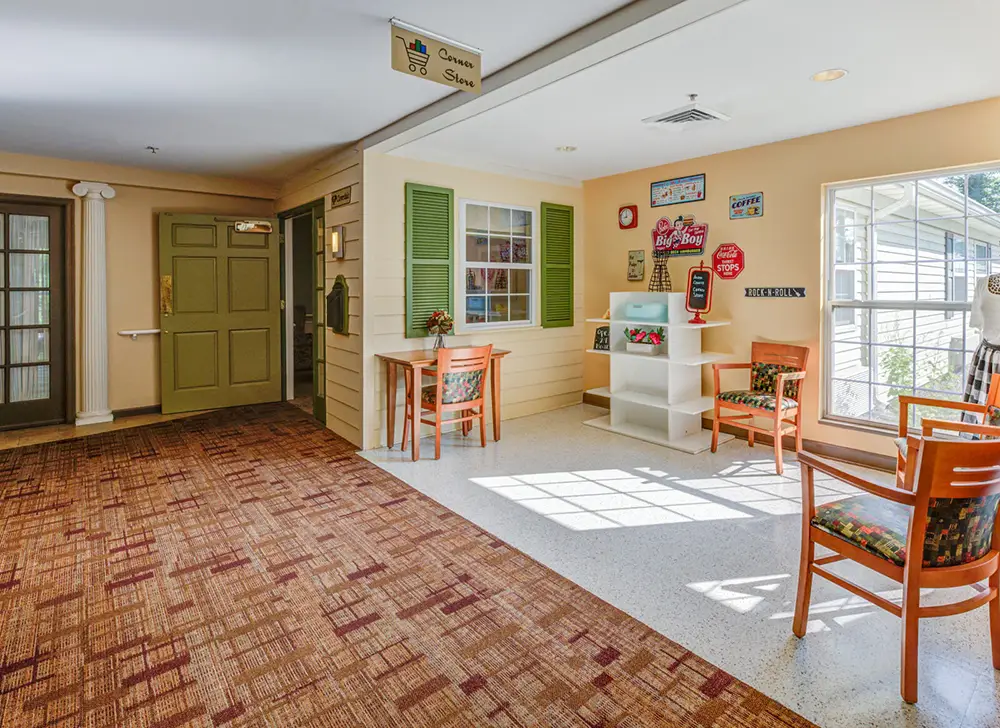 Allentown31 A small seating area with two wooden chairs and a table, wall shelves with decorations, and a window letting in natural light near a green door.