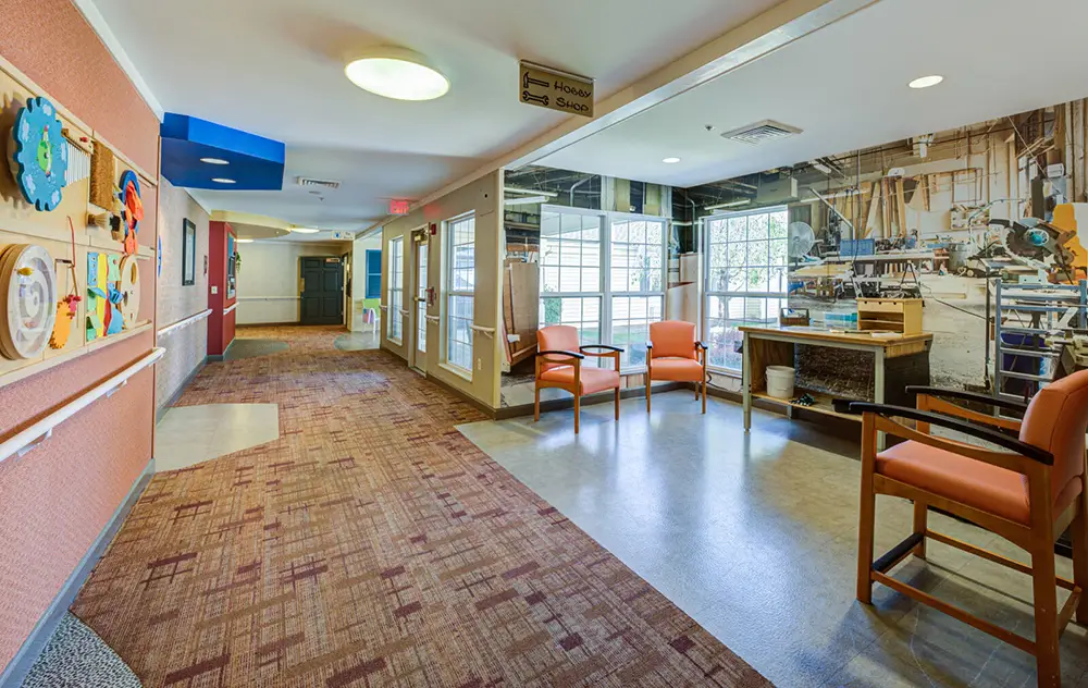 Allentown34 A hallway with patterned carpet, orange chairs, wall art, and large windows letting in natural light; a play area and equipment room are visible.