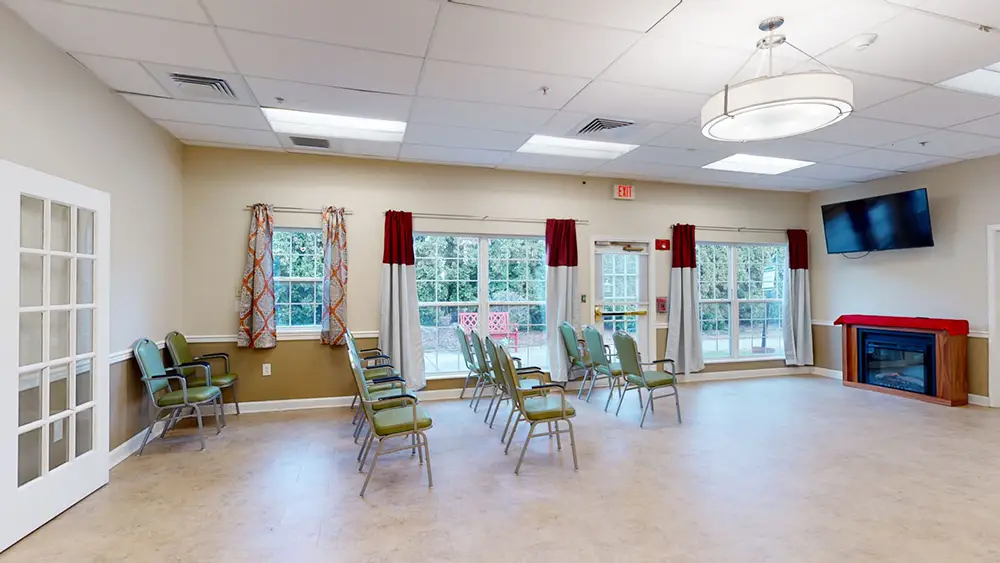 Allentown35 A spacious room with green chairs arranged in rows facing a wall-mounted TV, large windows with curtains, a fireplace, and French doors leading outside.