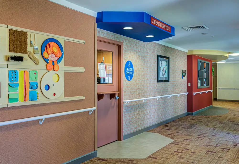 Allentown37 A hallway in a care facility with a sensory activity board on the wall, a door labeled "Health Center," and carpeted flooring.