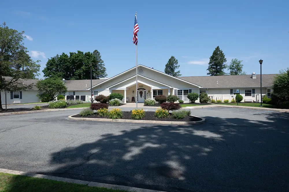 Allentown39 Single-story building with a covered entrance and American flagpole in front, surrounded by landscaping and a circular driveway under a clear blue sky.