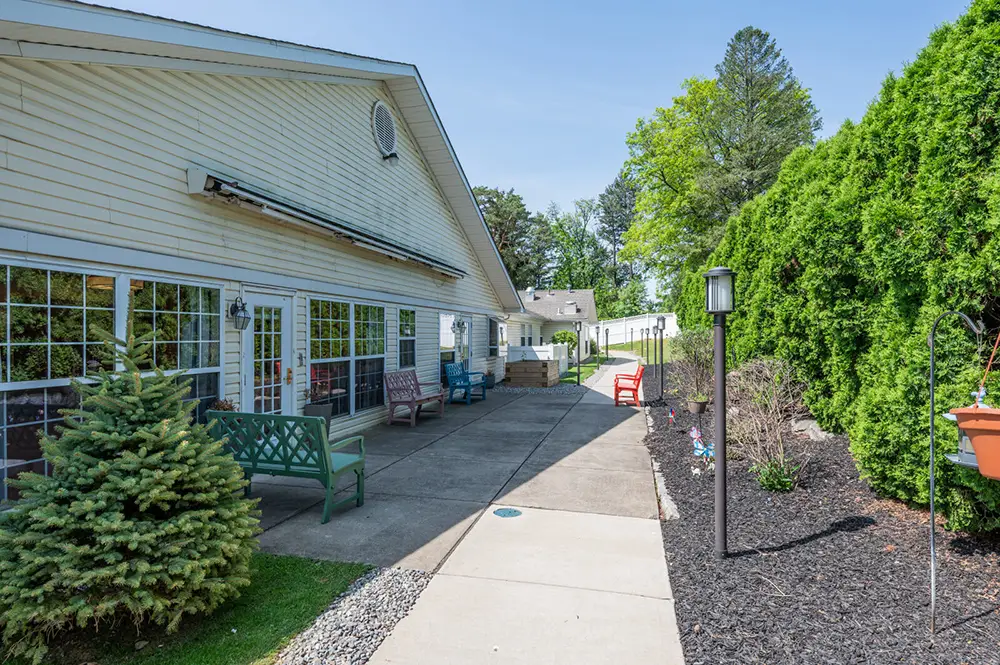 Allentown4 A paved walkway with benches runs alongside a building and landscaped greenery, with trees and a clear sky in the background.
