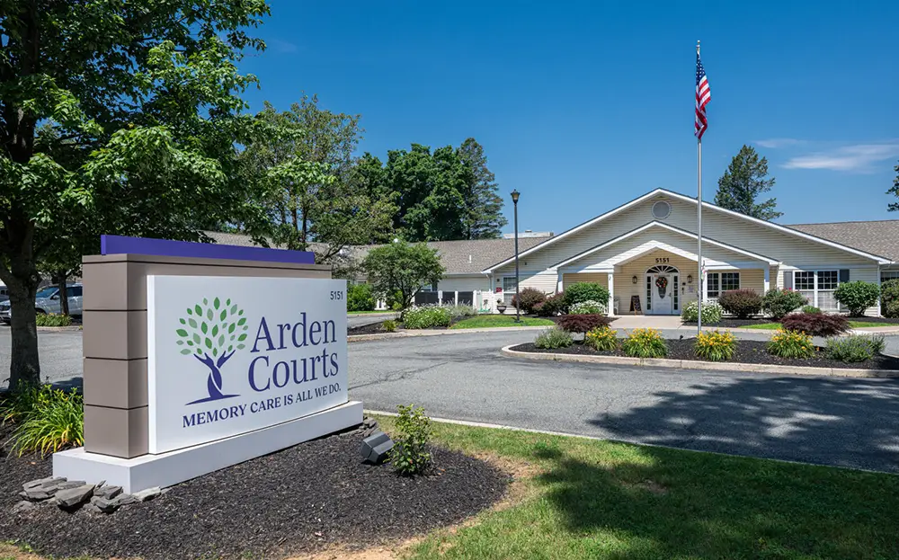 Allentown40 Arden Courts memory care facility exterior with sign in foreground, landscaped grounds, and American flag near entrance under a clear blue sky.