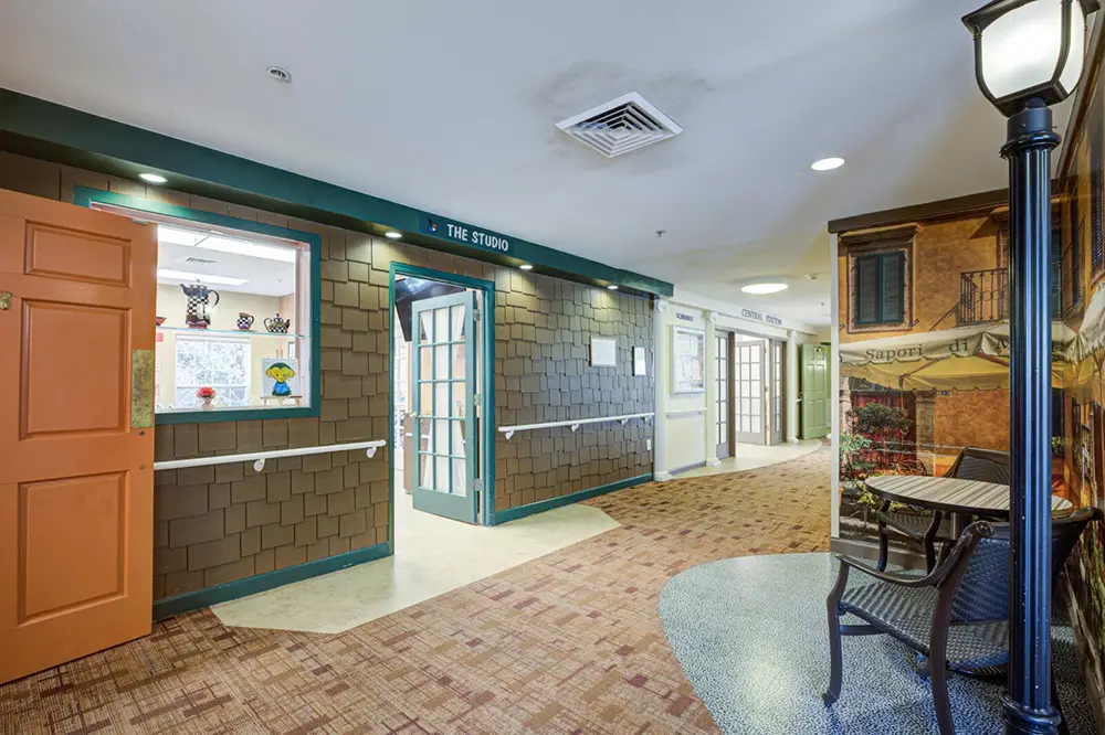 Allentown5 Indoor hallway with themed decor, featuring shop-like doors, a streetlamp, a mural of a building, and a small table with two chairs next to the wall.