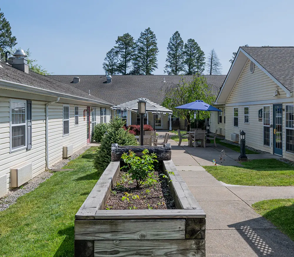 Allentown6 A courtyard with two single-story buildings, a raised garden bed, outdoor seating areas with umbrellas, and a sidewalk surrounded by grass and trees.