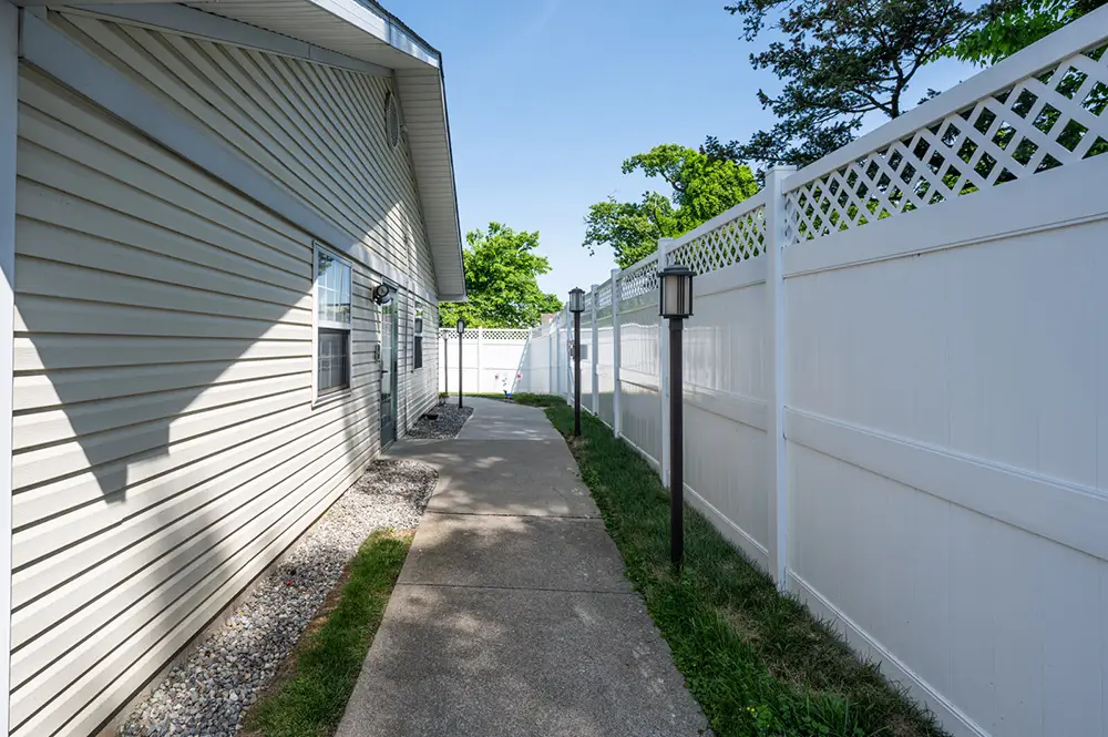 Allentown7 A concrete walkway runs alongside a beige house with vinyl siding, bordered by white privacy fencing and a lamp post, under a clear blue sky.