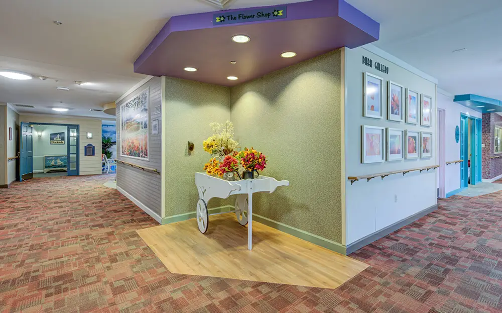 Allentown8 A white flower cart with artificial flowers is positioned under a purple “The Flower Shop” sign in a hallway with framed pictures on the wall and patterned carpet flooring.