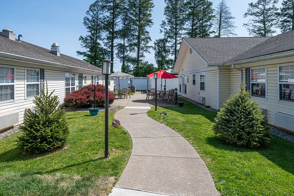 Allentown9 A concrete walkway curves between two single-story buildings, with grass, small trees, shrubs, and outdoor seating under a red umbrella.