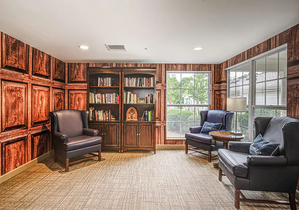 A cozy reading room with two navy armchairs, a wooden bookshelf filled with books, a small round table, and large windows letting in natural light. A cozy reading room with two navy armchairs, a wooden bookshelf filled with books, a small round table, and large windows letting in natural light.