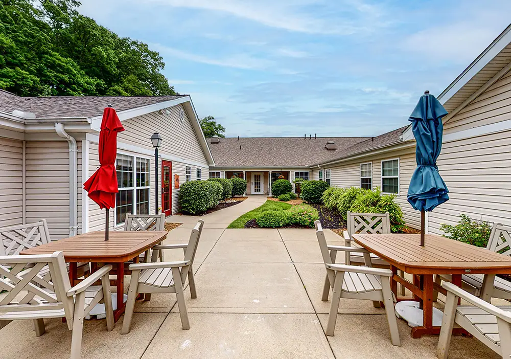 Outdoor patio area at Arden Courts in Anderson Township features wooden tables, chairs, and closed red and blue umbrellas, surrounded by beige buildings and landscaped greenery—creating a peaceful setting for memory care residents under a partly cloudy sky.