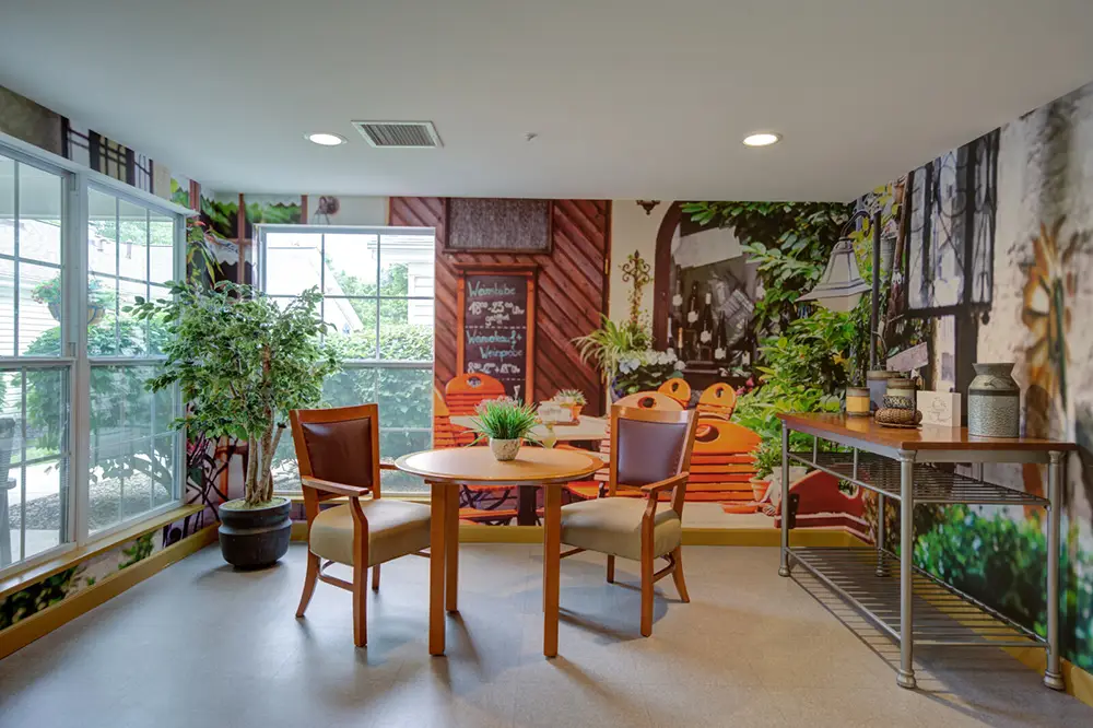 A small dining area with a round wooden table, two chairs, potted plants, a metal shelf, and a mural of a café scene on the wall. Large windows provide natural light.