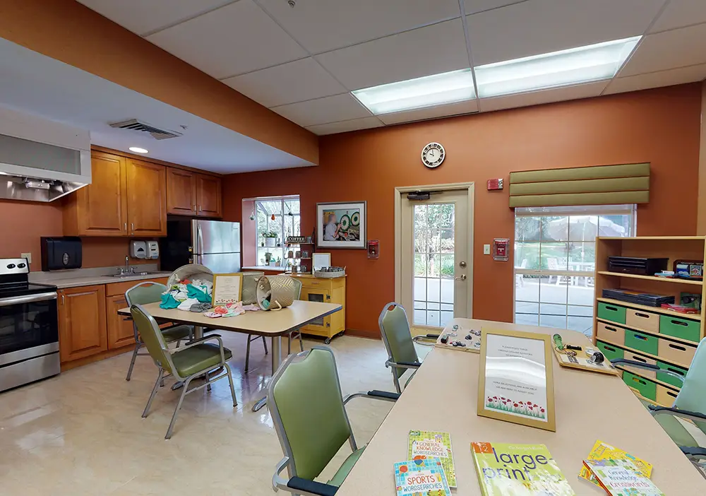A multipurpose room with a kitchen area, tables, chairs, storage bins, books, and large windows providing natural light. A multipurpose room with a kitchen area, tables, chairs, storage bins, books, and large windows providing natural light.