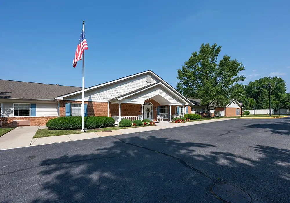 Single-story brick building with a white portico and an American flag out front, Arden Courts of Annandale offers specialized memory care, set against a blue sky and surrounded by green landscaping.