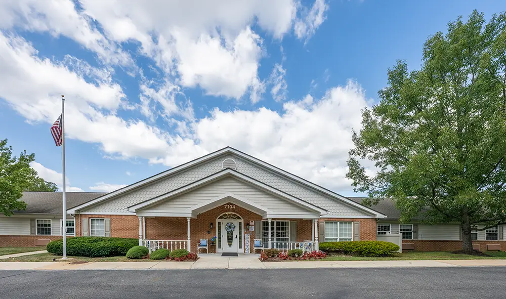 Single-story brick building with a white entrance porch, columns, and landscaping; an American flag is on a pole to the left and a large tree stands to the right at Arden Courts, a memory care community in Annandale.