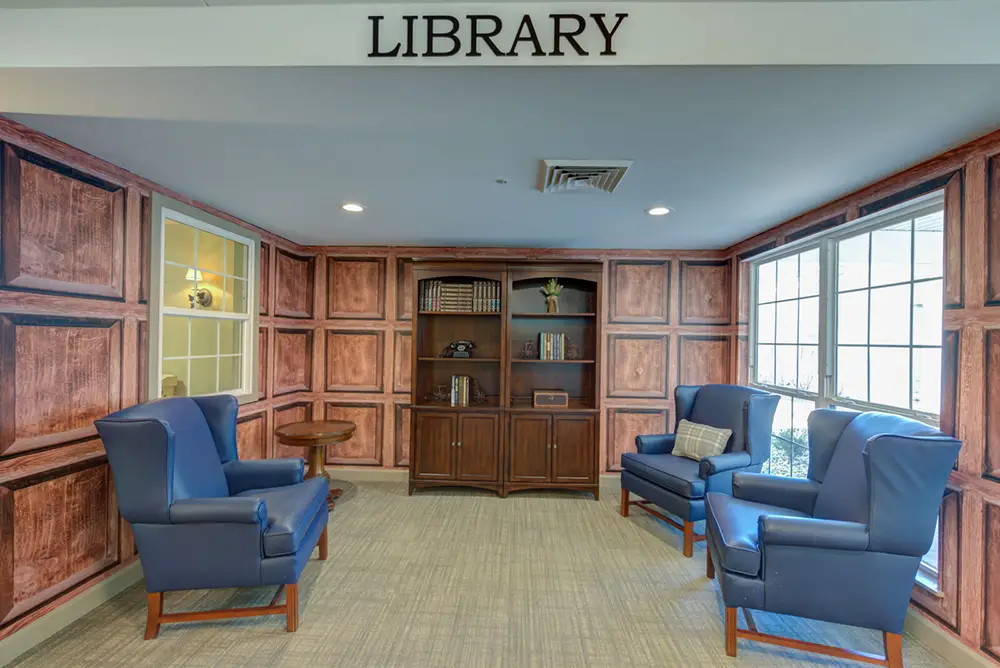 A small library room with four blue armchairs, a wooden bookshelf with books and decor, wood-paneled walls, and a large window letting in natural light.