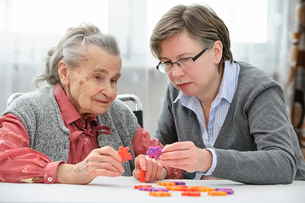 An elderly woman and a younger woman sit at a table, working together to assemble a colorful puzzle.