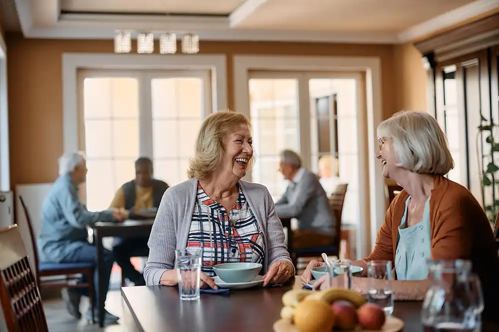 Two older women sit at a table, smiling and talking over a meal, with other seniors seated at tables in the background in a well-lit dining room.