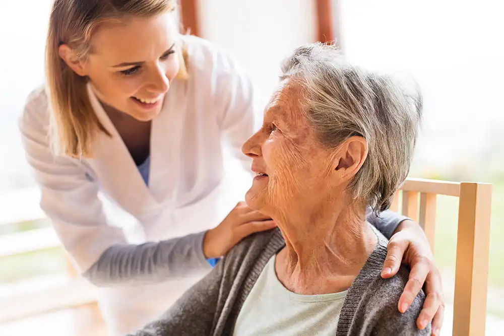 A young caregiver smiles and places a reassuring hand on the shoulder of an elderly woman sitting in a chair, both looking at each other.