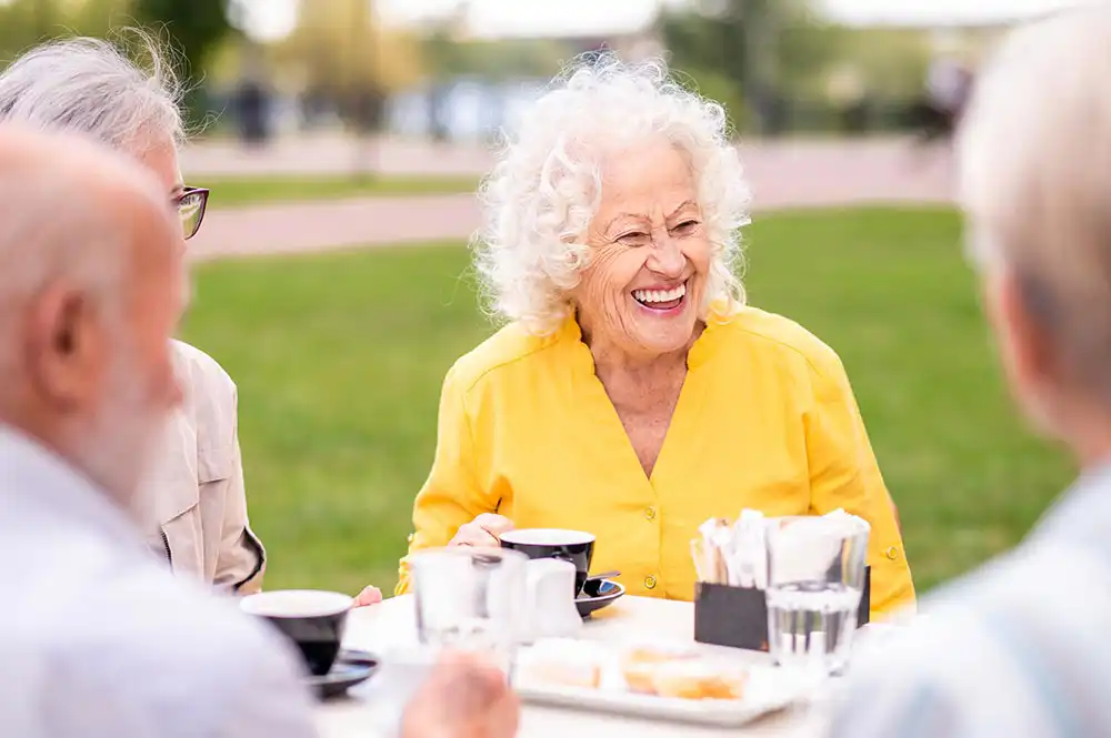 Elderly woman in a bright yellow shirt smiling while sitting outdoors at a table with others, drinking coffee and eating pastries.