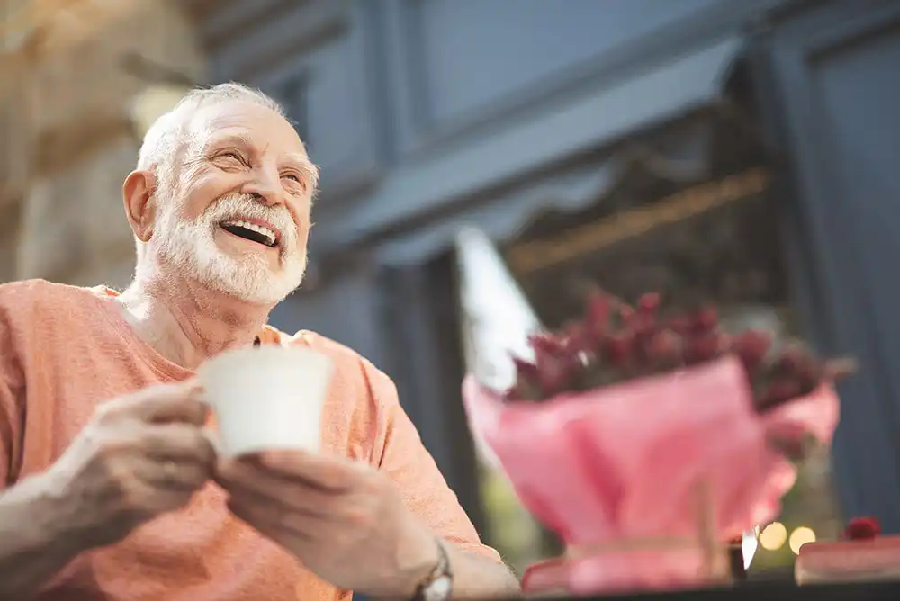 An older man with a white beard smiles while holding a white teacup, seated at a table with a pink-wrapped bouquet of flowers in the foreground. An older man with a white beard smiles while holding a white teacup, seated at a table with a pink-wrapped bouquet of flowers in the foreground.
